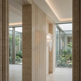A modern hallway features light stone walls, vertical textured panels, the Aeris 3 Light Wall Light in white, and large windows with views of a lush minimalist courtyard garden filled with assorted green plants.