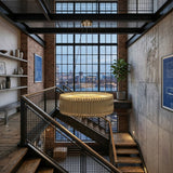 A modern industrial loft features large grid windows, exposed brick walls, metal stairs, shelves with ceramics, a potted plant, city skyline and bridge views outside, and is illuminated by the Aurelio 24 Light Brass Chandelier - Gold.