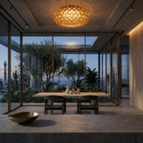 Modern dining area with large glass walls, a wooden table and chairs, the Briller LED Flush Ceiling Light in gold, and an outdoor terrace featuring trees and cityscape views at dusk.