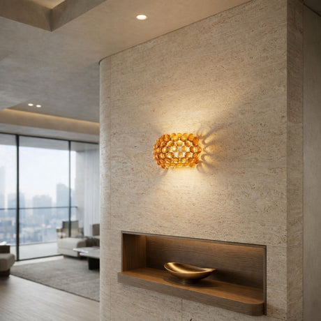 A modern living room with large windows, a textured beige wall, a golden decorative bowl on a built-in shelf, and the Briller LED Small Wall Light - Gold casting amber patterned ambient lighting.