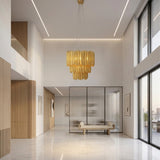 A modern, spacious lobby with high ceilings, large windows showing a cityscape, marble floors, wood accents, minimalist art, a beige bench, and a glass-walled office features the Aurelio 42 Light Brass Chandelier - Gold as its sculptural centerpiece.