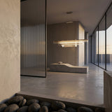 A modern minimalist interior with concrete floors, a sculptural bench, large glass window, and decorative black stones is enhanced by warm natural light and the Celestri LED Linear Suspension Light - Silver.