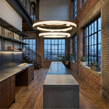 A modern industrial loft kitchen with tall windows, exposed brick walls, wood floors, a long concrete island, metal shelving, potted plants on the windowsills, and the gold Celine LED 2 Tier Suspension Light adding luxury lighting.