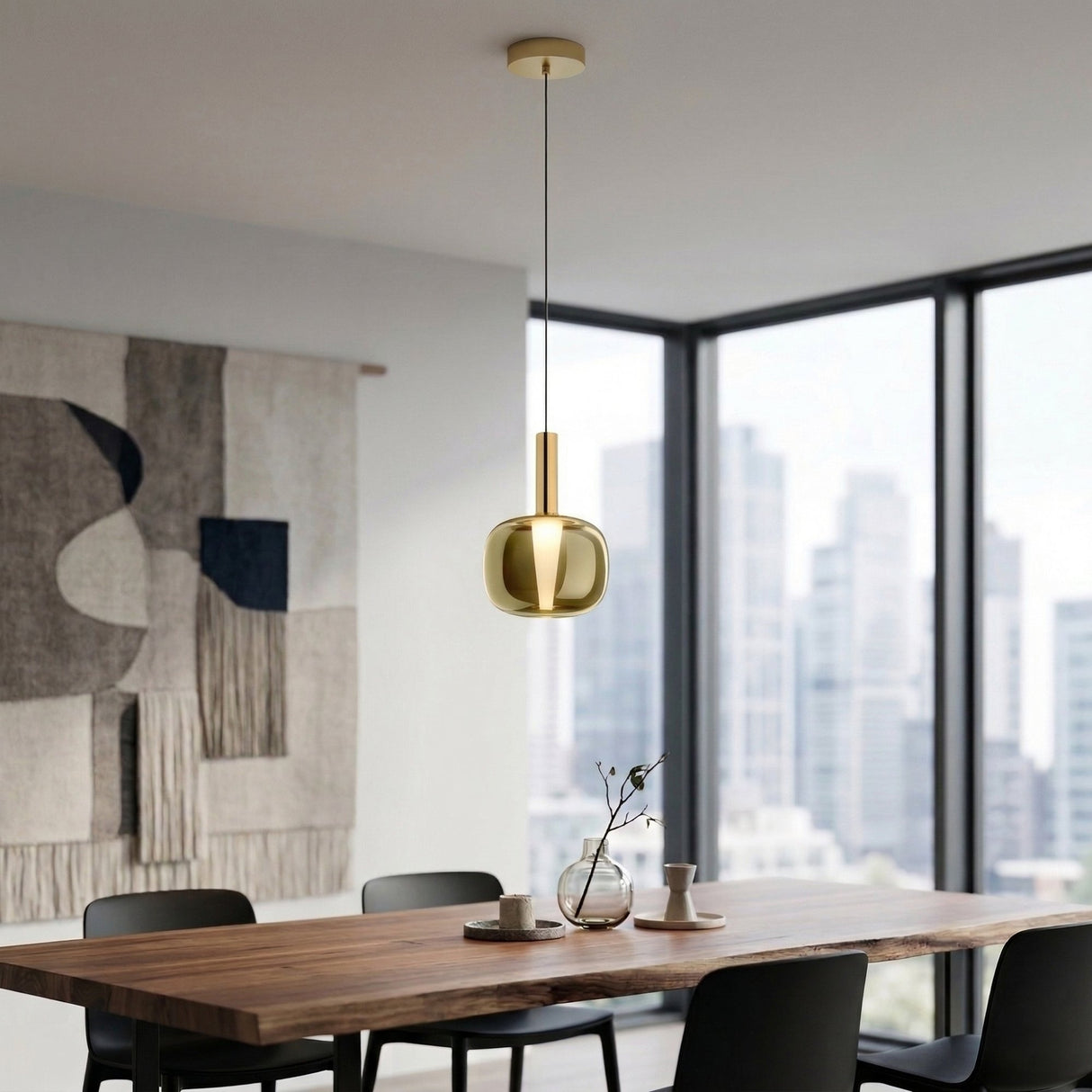 A modern dining room features a wooden table, black chairs, the Dusk Dawn LED Pendant Light in gold, a minimalist wall tapestry, large windows with a city view, and a small vase with branches on the table.