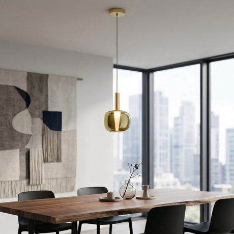 A modern dining room features a wooden table, black chairs, the Dusk Dawn LED Pendant Light in gold, a minimalist wall tapestry, large windows with a city view, and a small vase with branches on the table.