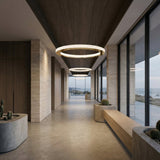 A modern hallway with large windows, wooden accents, and concrete planters holding cacti features the Celine LED Single Suspension Light in gold. Through the windows, a coastal landscape under a cloudy sky is visible.