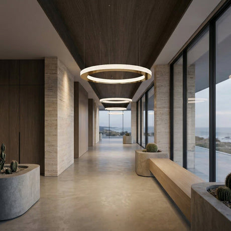 A modern hallway with large windows, wooden accents, and concrete planters holding cacti features the Celine LED Single Suspension Light in gold. Through the windows, a coastal landscape under a cloudy sky is visible.