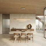 A modern dining room with minimalist design features a light wood table and four chairs, a sculptural centerpiece on a sideboard, neutral stone walls and floors, the Celestri LED Linear Suspension Light in silver, large windows, and city skyline views.
