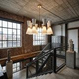 A modern industrial interior with exposed brick walls, large windows, concrete ceiling, black metal stair railings, lockers, and a Ramses 8 Light Chandelier - White with cone-shaped shades hanging above the stairs.