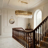 A grand staircase with wooden railings and marble floors, featuring the Royal 8-16 Light Crystal Chandelier in gold as a luxury lighting centerpiece, plus an arched window, round mirror, and a potted plant on a pedestal.