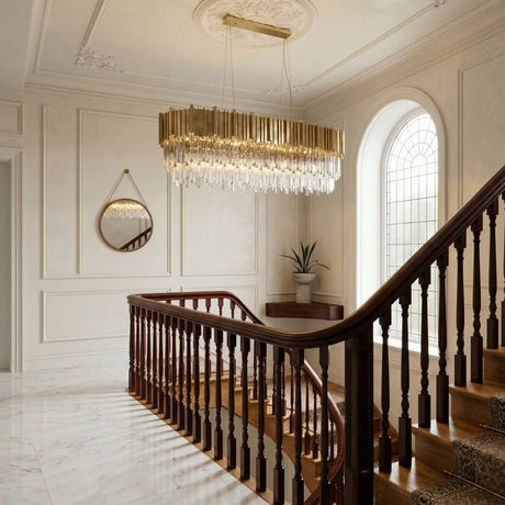A grand staircase with wooden railings and marble floors, featuring the Royal 8-16 Light Crystal Chandelier in gold as a luxury lighting centerpiece, plus an arched window, round mirror, and a potted plant on a pedestal.