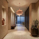A modern hallway with beige stone walls and floors, a wooden bench, abstract sculpture, large potted cactus, and the striking Picasso Pendant Light - Pink. Floor-to-ceiling windows at the end reveal a sweeping city view.