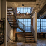 Niori Tree 7 Light Suspension in black with branching arms and clear glass globes, hanging in an industrial loft stairwell with brick walls and grid windows.