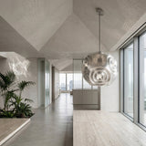 A modern, minimalist interior with light gray walls, large windows, a geometric ceiling, indoor plants, city skyline views, and the Space 1 Light Pendant - Silver as a striking Art Deco lighting feature.
