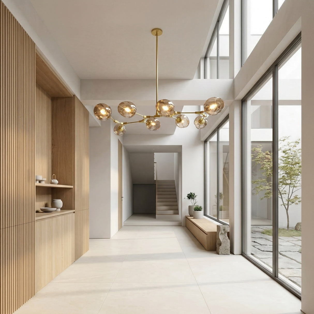 A modern, minimalist hallway features large windows, light wood accents, a Tree 8 Light Suspension - Gold chandelier, built-in shelving, a wooden bench, and views of a small courtyard with a tree.