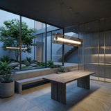 Niori Qbic LED Linear Suspension Light Small in clear glass and brass hangs above a wooden dining table in a modern room with a courtyard garden view.