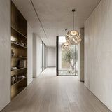 Minimalist hallway with light wood flooring, a Space 1 Light Pendant in silver, built-in gold shelves displaying pottery, and a glass door opening to a courtyard with a tree and white stones.
