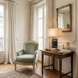 Elegant room with cream walls, tall windows, a vintage green-upholstered armchair, wooden table topped with the Caviron 1 Light Block Marble Table Lamp - Brass & Black, books, ceramics, framed mirror above, all on herringbone wood floor.