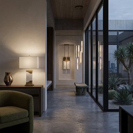 A modern interior hallway with a concrete floor, large windows, contemporary lighting, minimalist art, a green chair, the Torvani 1 Light Statement Marble Table Lamp in Brass & White, and desert plants visible outside in a courtyard.