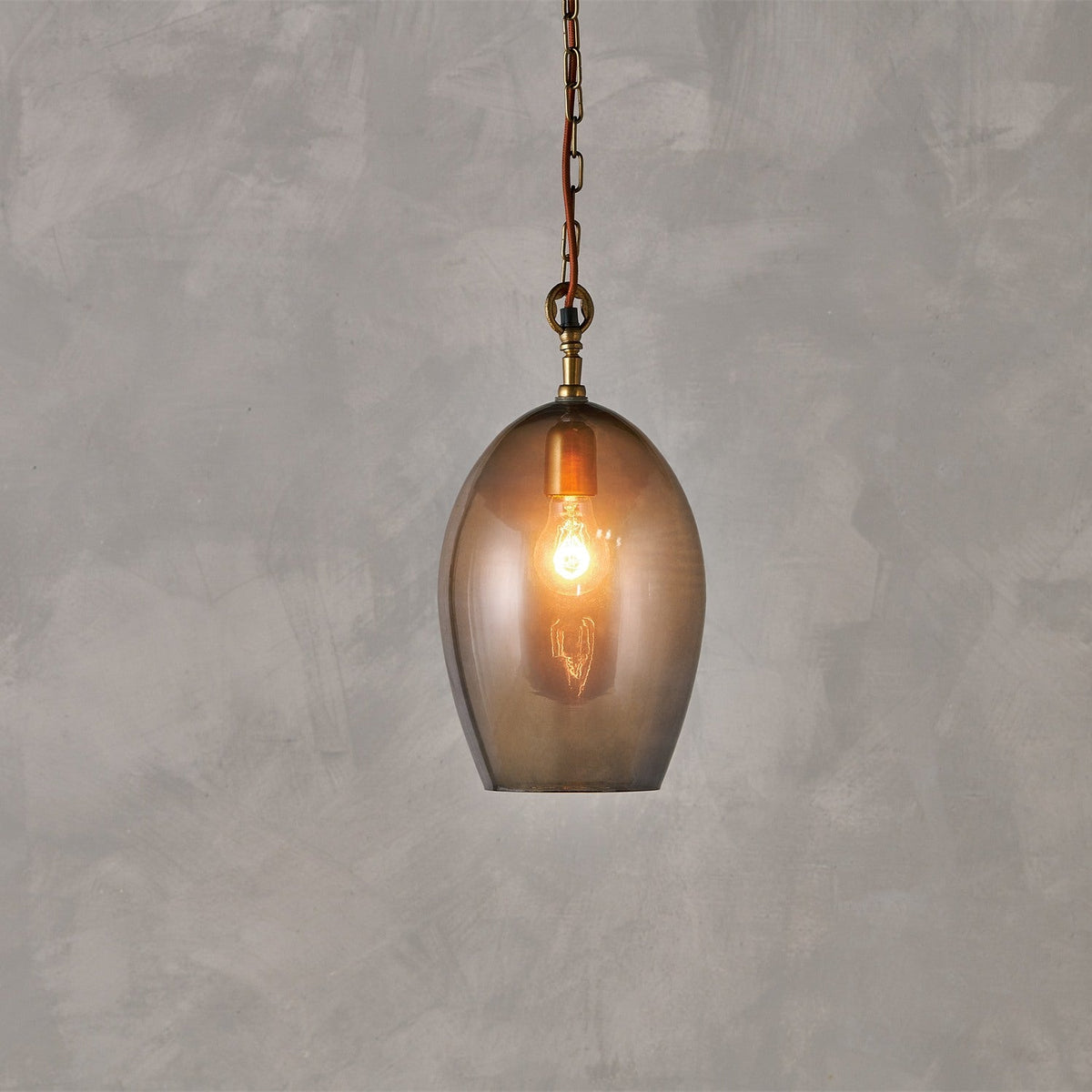 Otoro Small Oval Recycled Glass Pendant Light in Smoke, an egg-shaped glass shade with brass collar, ceiling rose, and chain, rust fabric flex, lit bulb visible, on a plain grey background.