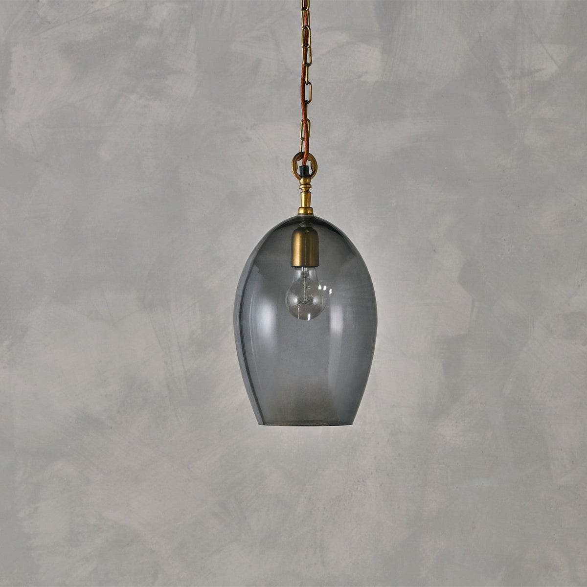 Otoro Small Oval Recycled Glass Pendant Light in smoke grey, with a brass collar and ceiling rose, rust fabric flex and chain, exposed bulb, on a plain grey background.