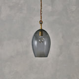 Otoro Small Oval Recycled Glass Pendant Light in smoke grey, with a brass collar and ceiling rose, rust fabric flex and chain, exposed bulb, on a plain grey background.