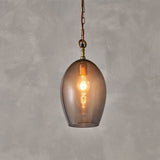 Otoro Small Oval Recycled Glass Pendant Light in Smoke, an egg-shaped glass shade with brass collar and chain on a rust fabric flex, shown lit against a plain grey background.