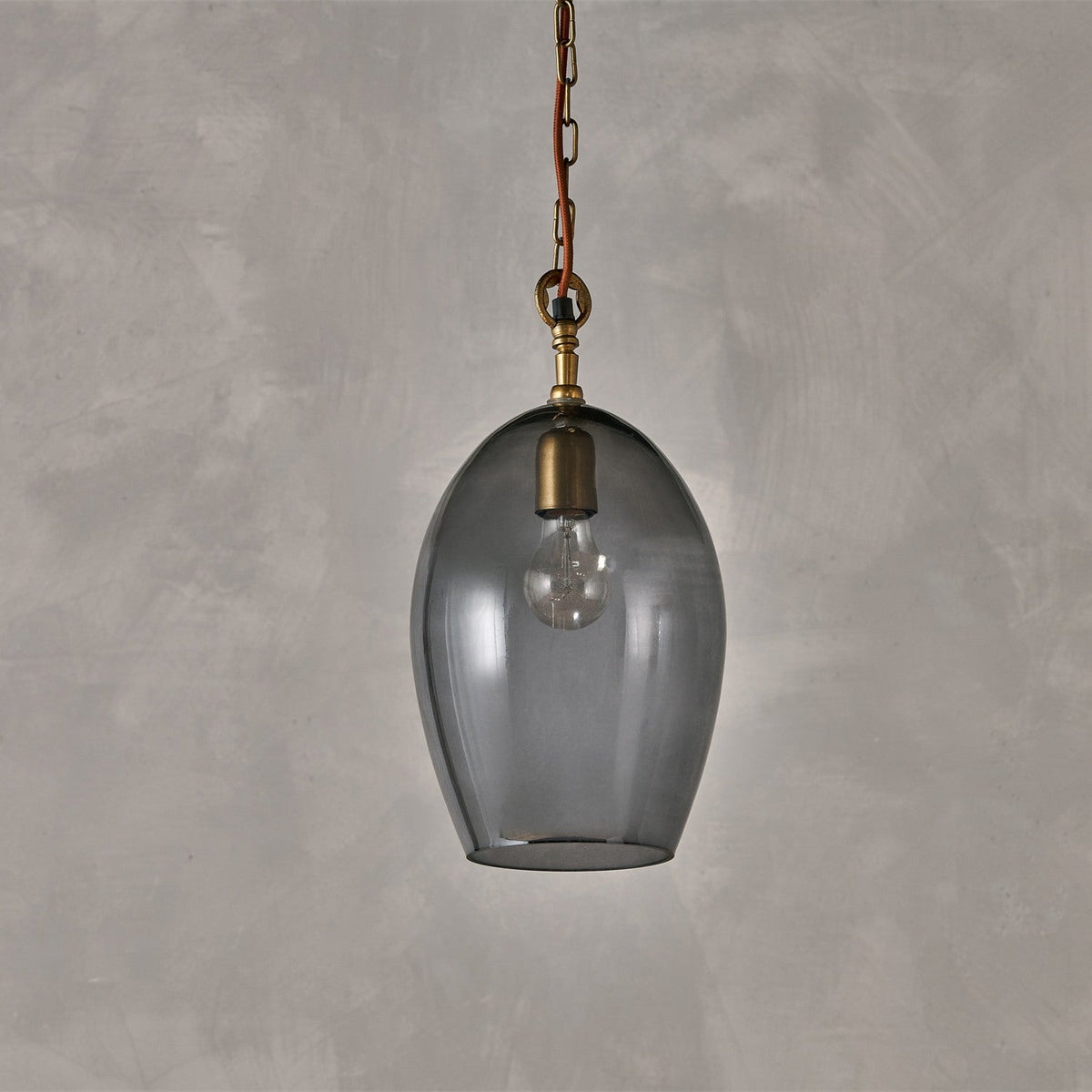 Otoro Small Oval Recycled Glass Pendant Light in smoke, an egg-shaped glass shade with brass collar and chain on rust fabric flex, shown against a plain grey background.