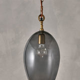 Otoro Small Oval Recycled Glass Pendant Light in smoke, an egg-shaped glass shade with brass collar and chain, exposed bulb visible, shown against a plain grey background.