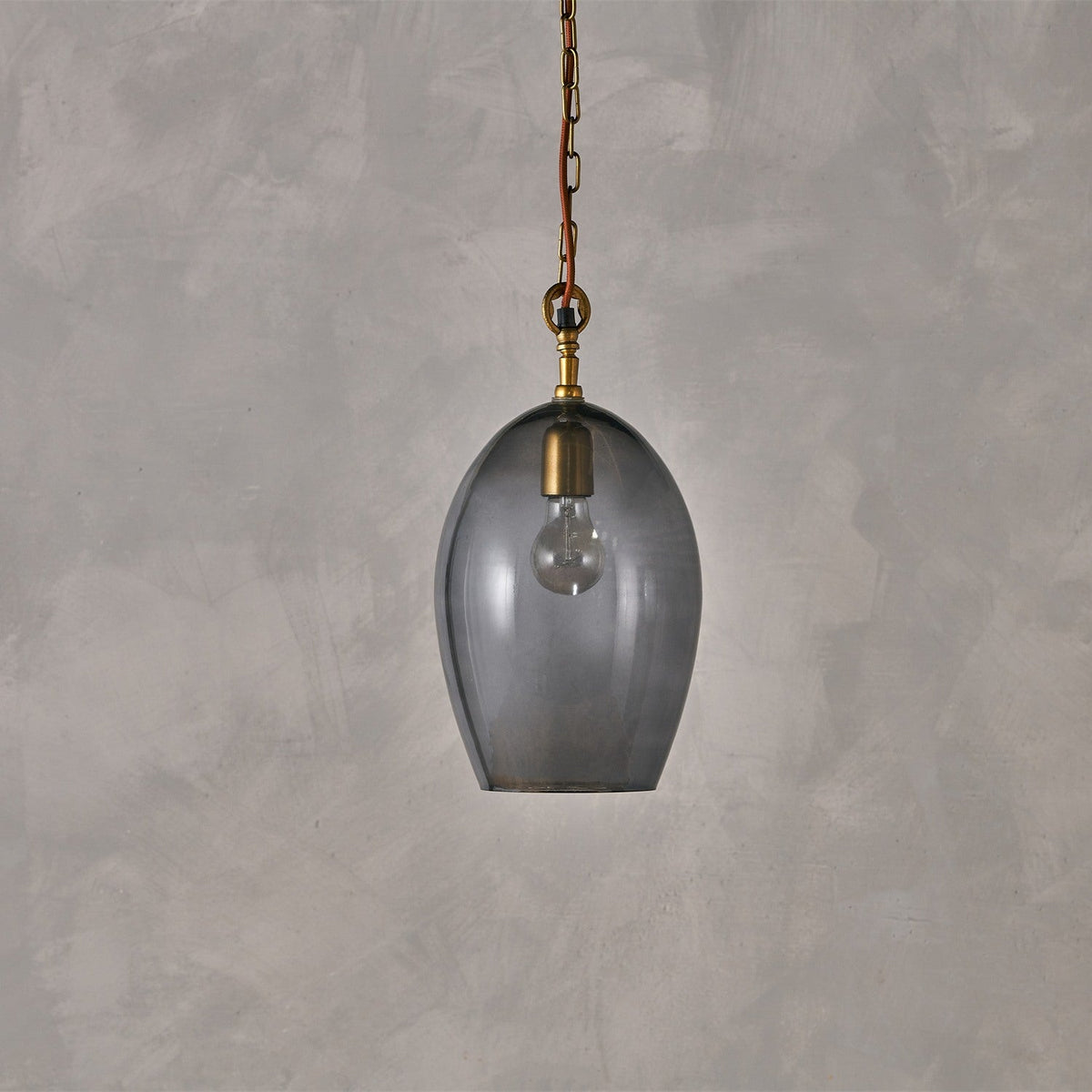 Otoro Large Oval Recycled Glass Pendant Light in smoke, an egg-shaped shade with brass collar and chain on rust fabric flex, shown against a plain grey background.