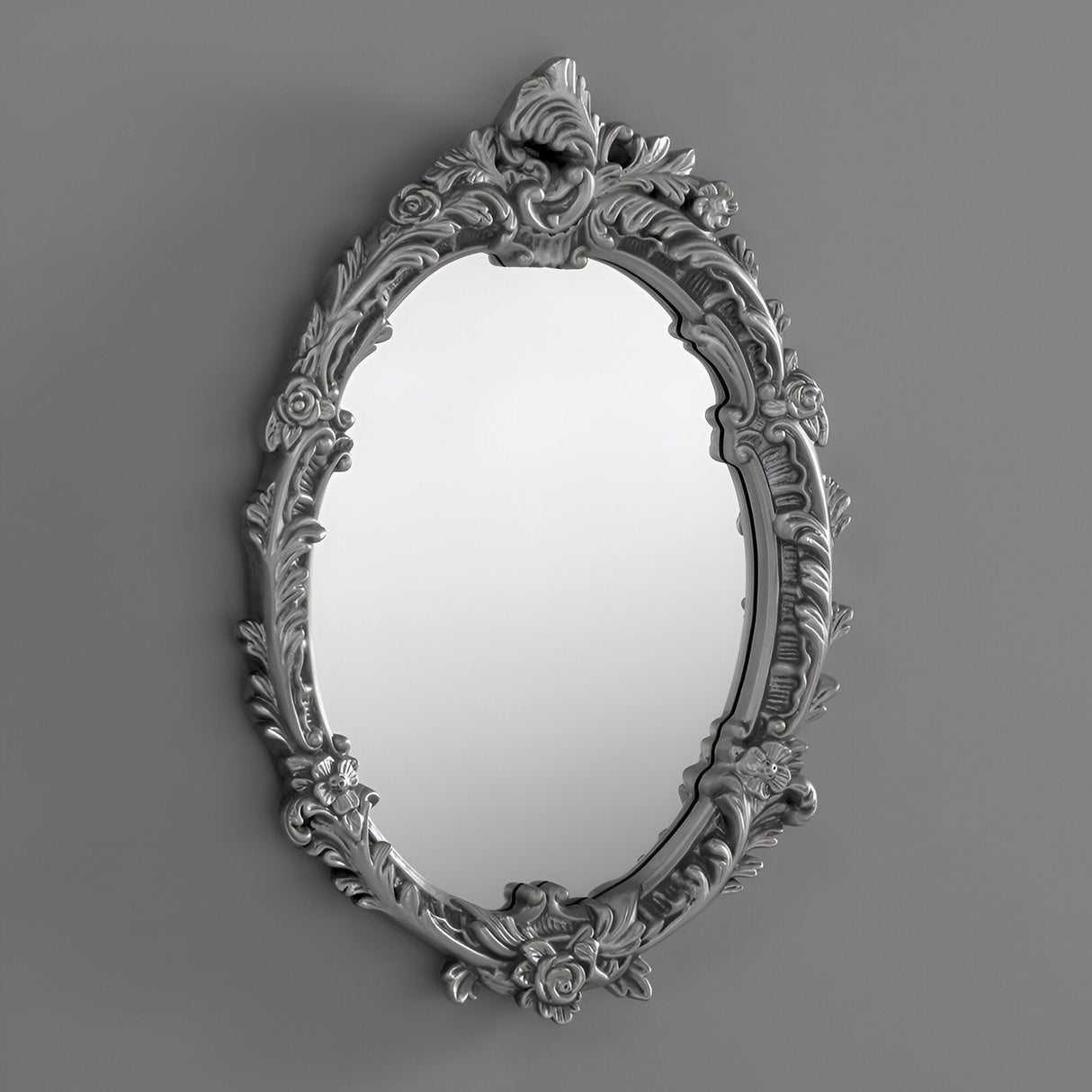 The Illuna Oval Wall Mirror Silver - 64x46cm features an ornate floral design and a metallic silver finish, effortlessly complementing diverse interior styles against a simple gray backdrop.