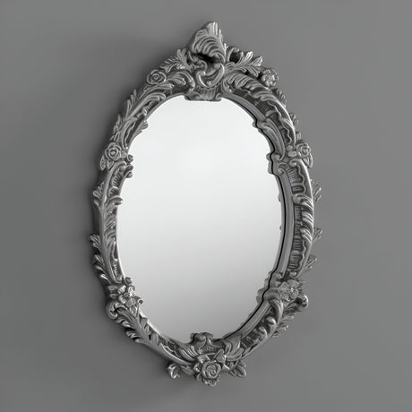 The Illuna Oval Wall Mirror Silver - 64x46cm features an ornate floral design and a metallic silver finish, effortlessly complementing diverse interior styles against a simple gray backdrop.