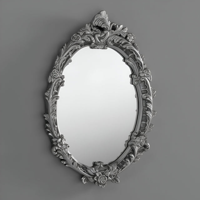 The Illuna Oval Wall Mirror Silver - 64x46cm features an ornate floral design and a metallic silver finish, effortlessly complementing diverse interior styles against a simple gray backdrop.