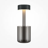 The AI Collaboration Rechargeable LED Table Lamp features a smoked glass vase-style base, cylindrical design, and a flat round black shade. Its translucent stem provides soft, warm light and enhances its minimalist elegance against plain backgrounds.