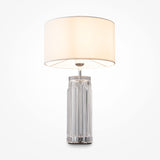 The Muse Table Lamp - Chrome With White Shade features a cylindrical, transparent glass base with vertical ridges, ideal for modern interiors. A wide, cream-colored fabric shade emits a warm glow and is elegantly set against a plain white background.