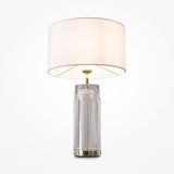 The Muse Table Lamp - Gold With White Shade boasts a modern design with a cylindrical glass base and subtle gold accents, topped with a large white fabric shade. It stands elegantly against a white backdrop, seamlessly blending style and simplicity.
