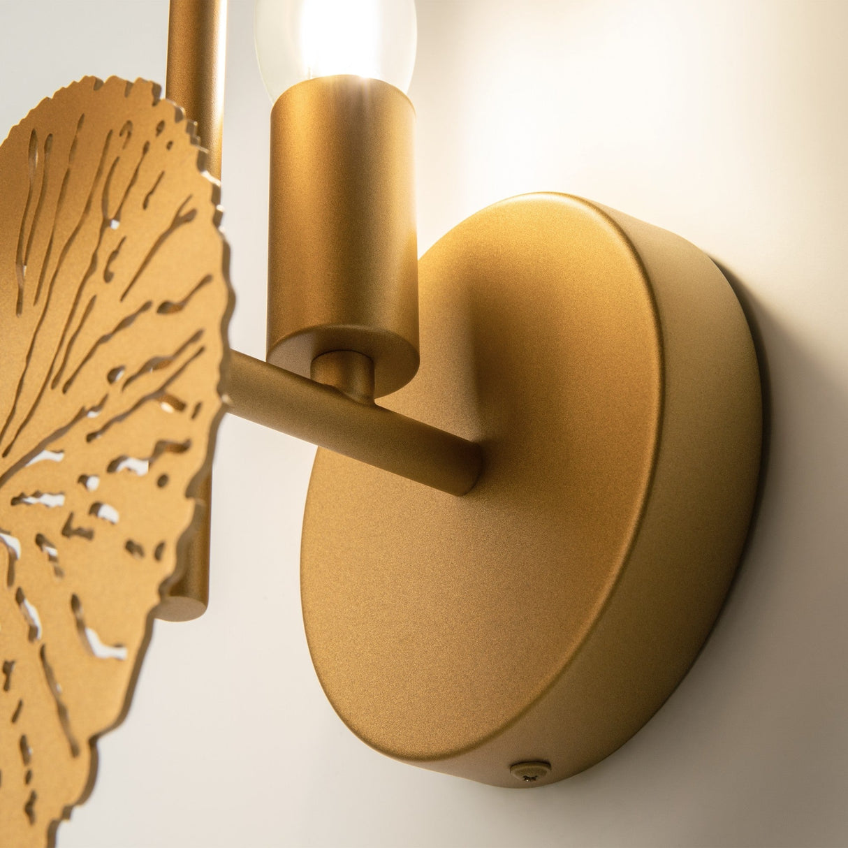 Close-up of the Lovetann Wall Light - Gold With Perforated Metal Discs. The modern sconce has a round gold base and a subtle floral-inspired design on the left, with its sleek form illuminated by a bulb at the top.