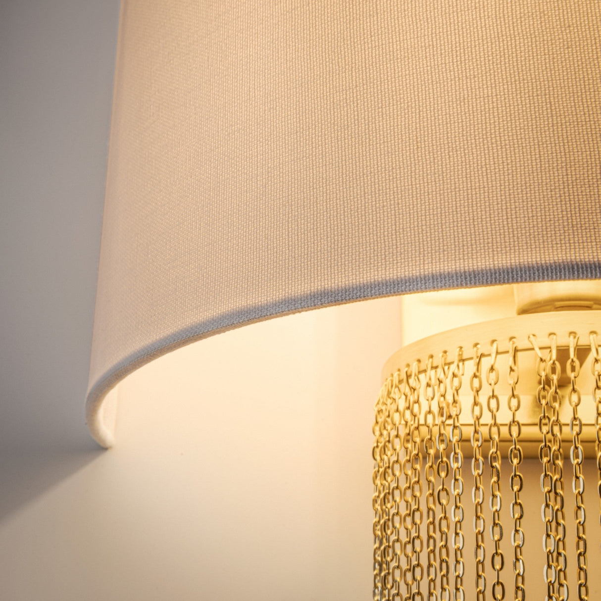 Close-up of the Impressive Wall Light, featuring a beige fabric shade and a base adorned with hanging gold chains. The ambient lighting softly highlights the material textures.