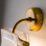 A close-up of the Cabaret LED Curved Wall Light - Brass showcases its modern design, with a curved arm and a transparent rectangular glass shade. It features a smooth circular base that elegantly matches the light-colored wall background.