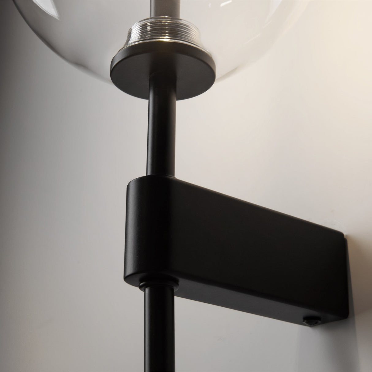 A close-up shows the Mood Wall Light - Black With Clear Round Glass, featuring a sleek cylindrical black metal structure and a clear glass globe encasing the bulb, mounted on a light-colored wall for a minimalist design.