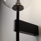 A close-up shows the Mood Wall Light - Black With Clear Round Glass, featuring a sleek cylindrical black metal structure and a clear glass globe encasing the bulb, mounted on a light-colored wall for a minimalist design.