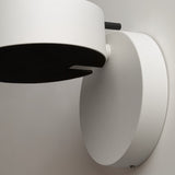 The Nuance LED Movable Head Wall Light in white with black detail features a circular base and cylindrical lamp, delivering sleek modern lighting with energy-saving LED technology when mounted on a light-colored wall.