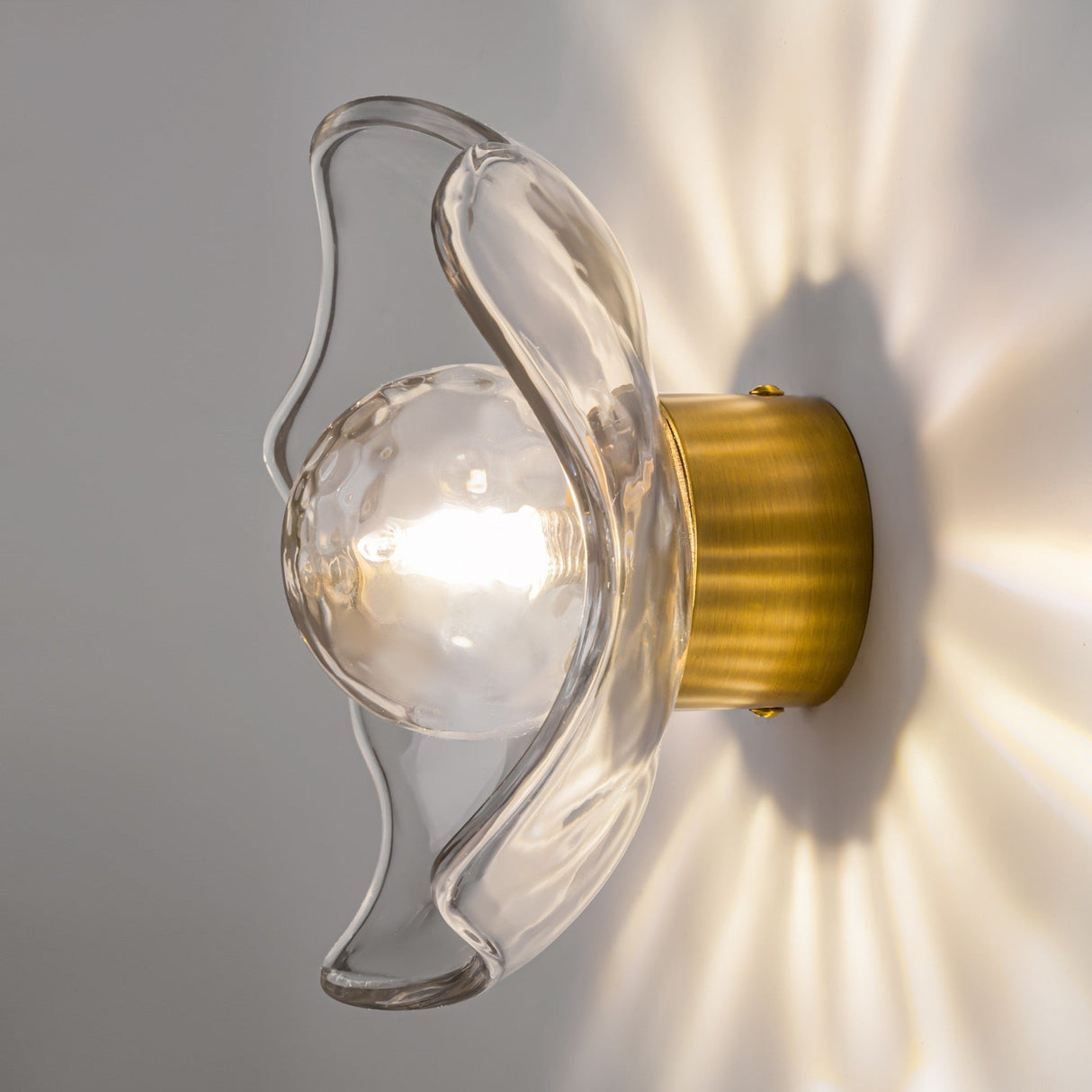 The Miracle Single Wall Light in brass boasts a clear, flower-shaped cast glass shade, housing a glowing bulb. Its elegant Art Deco design casts wavy patterns on the wall, adding vintage sophistication to any room.