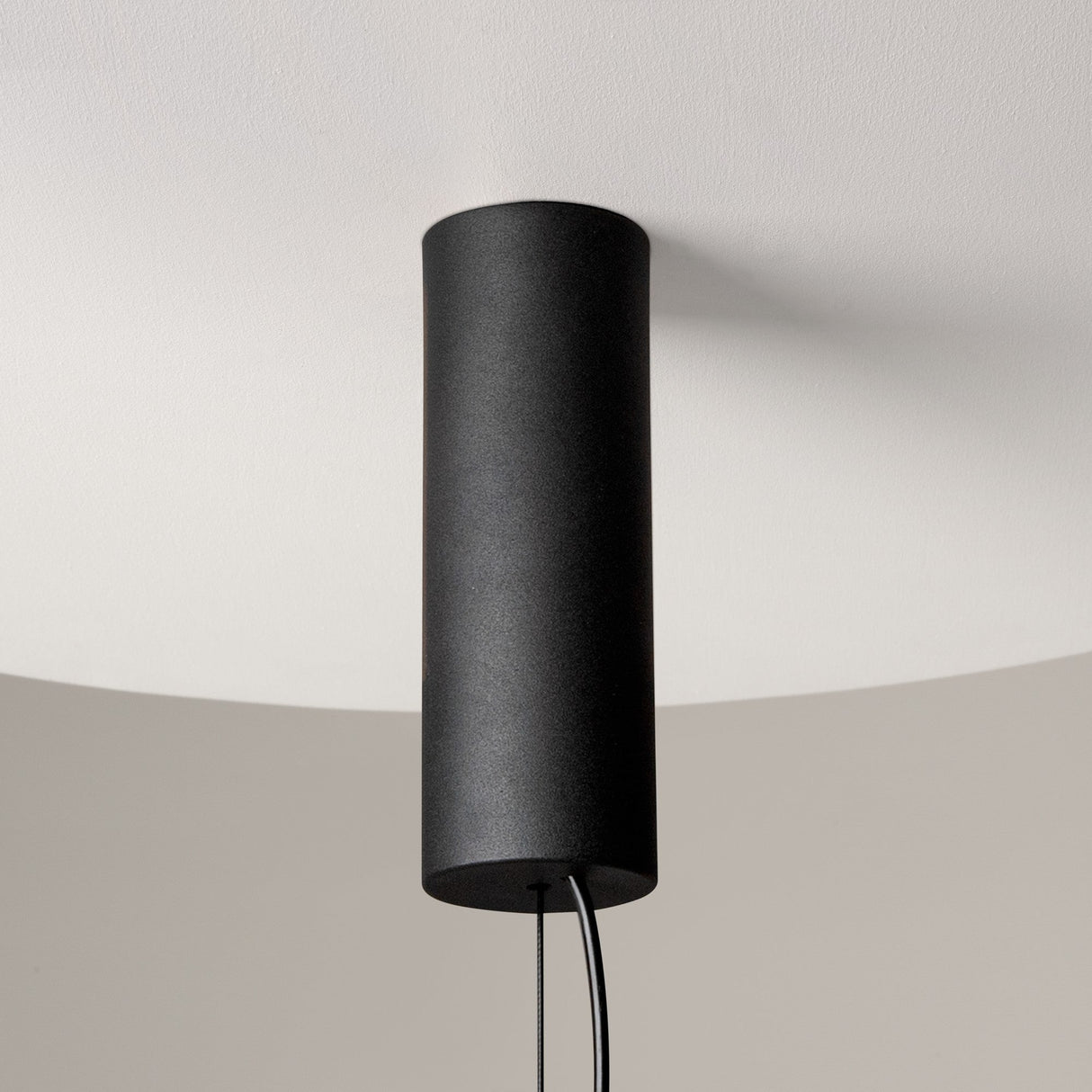 The Cosmo LED Linear Suspension Light, featuring a cylindrical black design with clear glass elements, hangs from a white ceiling with its cord visible at the bottom, offering a futuristic look that contrasts elegantly against the softly lit background.