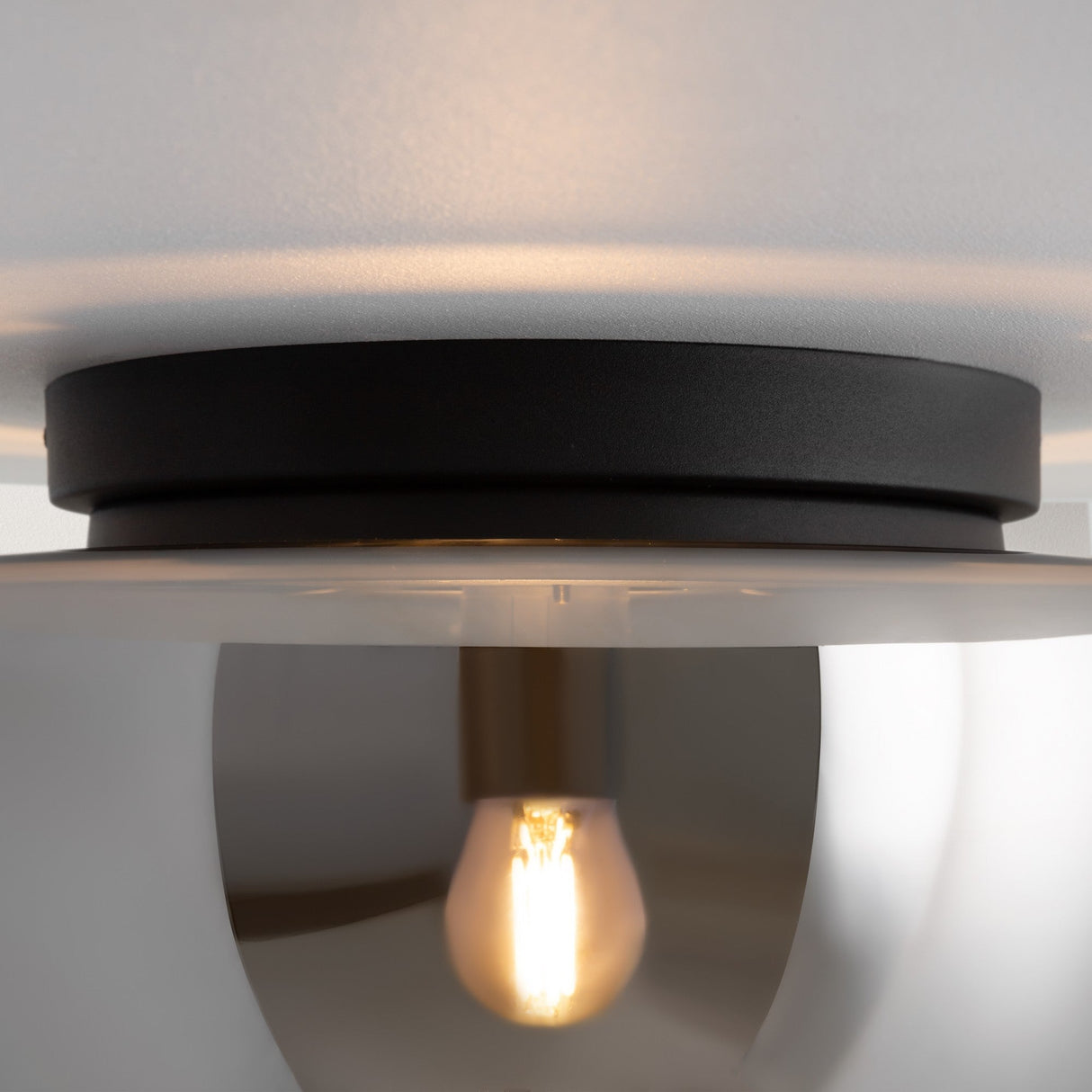 The Glassy Ceiling Flush Light features an exposed bulb within a reflective metallic dome, ideal for compact spaces. It casts a warm glow through smoky black glass and is mounted on a white ceiling.