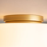 The Glassy Ceiling Flush Light features a round, gold-colored metal base with white matte glass that evenly diffuses light. Its low-profile design fits snugly against the ceiling, highlighting its metallic sheen.