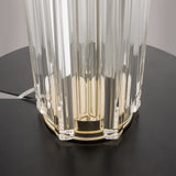 Close-up of the Muse Table Lamp - Gold With White Shade, showcasing its vertical translucent glass panels and gold-toned base. The modern piece sits elegantly on a round black table with the power cord visible behind it.