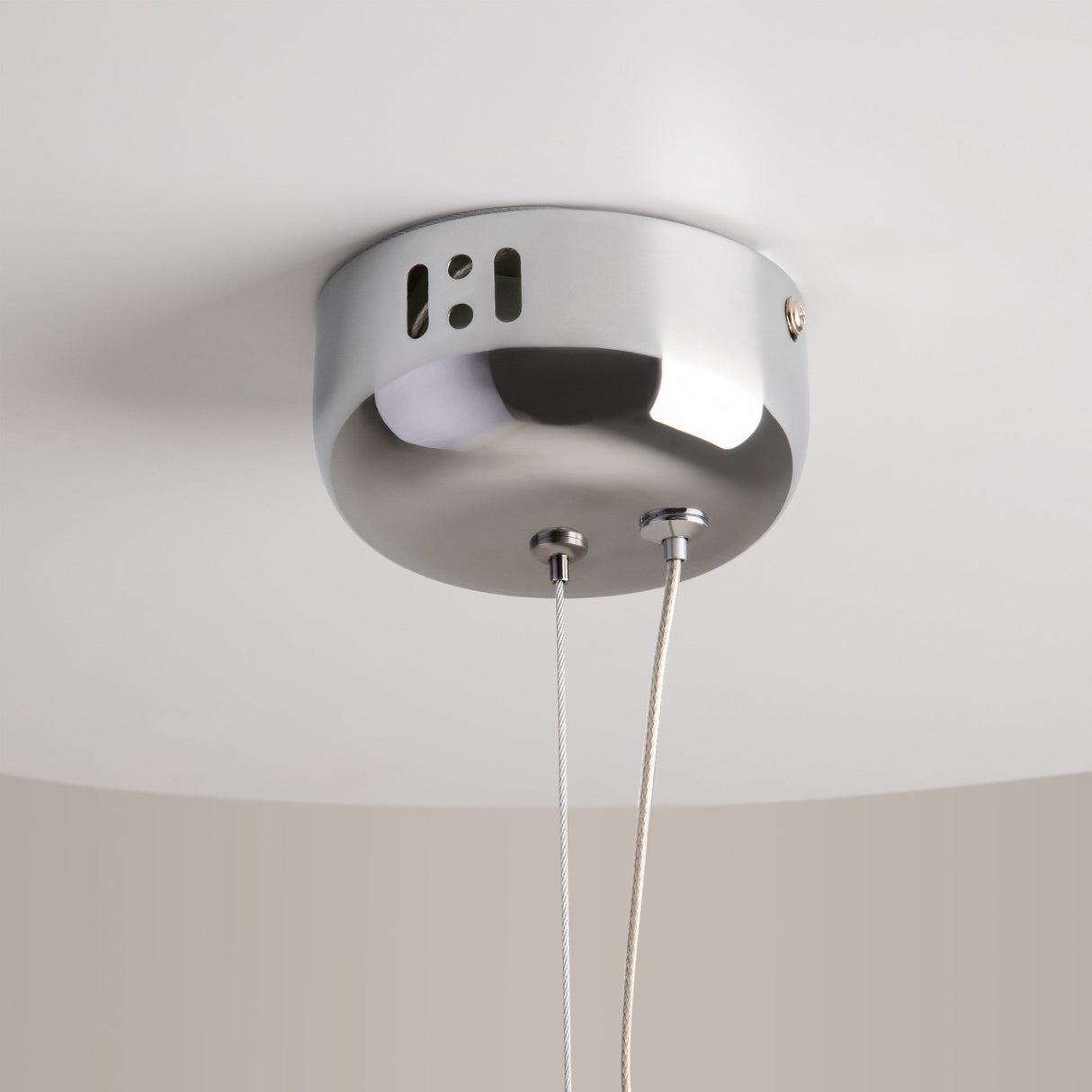 The Rebel LED Glass Pendant Light - Chrome features a sleek, circular design with a smooth, reflective surface and small rectangular cutouts. It hangs from two slender cables on a white ceiling.