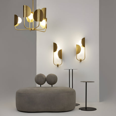 Minimalist room with a gray cushioned chair, two black metal side tables, and a modern chandelier. Two Tropic 2 Light Right High Wall Lights in brass feature circular lights partially covered by textured gold panels—a luxurious tropical getaway touch.
