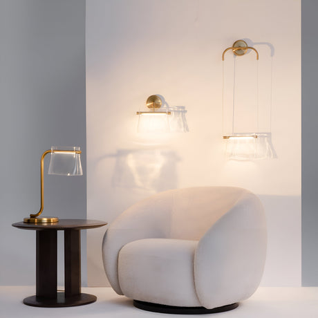 A cozy interior features a rounded white armchair beside a small round wooden side table. The space is illuminated by two Cabaret LED Curved Wall Lights in brass, with transparent glass shades that cast warm light on the wall and furniture.